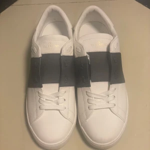 Valentino Garavani snörade sneakers vit/svart - Snygga Valentino Garavani snörade sneakers i vitt skinn med bred mörkblå läderdetalj över vristen. Klassisk rund tå, vita snören och coola nitar baktill på sulan. Perfekta för dig som gillar stilrena men ändå unika skor. Pris kan diskuteras kommer tyvvär utan kvitto.