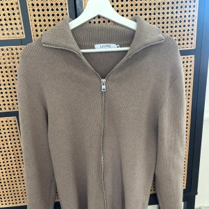 Beige ribbad kofta från Ljung - Snygg beige ribbad kofta från Ljung med hel dragkedja och hög krage. Tröjan är långärmad och har en clean, stilren look som passar perfekt till jeans eller skjorta. Materialet är stickat och känns både mjukt och bekvämt.