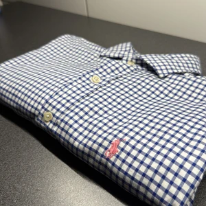 Blårutig skjorta Polo Ralph Lauren M - Klassisk blårutig skjorta från Polo Ralph Lauren i slim fit. Skjortan har vit botten med blå rutor, knappar framtill och broderad rosa logga på bröstet. Tillverkad i mjuk bomull och har långa ärmar med manschettknappar. Perfekt för en clean och stilren look.