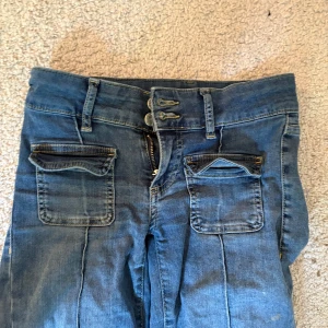 Blå bootcut jeans från Nelly - Snygga blå jeans från Nelly med bootcut passform och låg midja. Jeansen har två stora fickor framtill med lock och dubbla knappar i midjan. Tillverkade i klassiskt denimtyg och har markerade sömmar längs benen för en cool look. Den ända defekten är att de spilta lite glitter på ena benet som knappt syns 