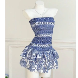 Blåvit strapless klänning - Supersöt blåvit kortklänning med strapless design och smockad överdel. Kjolen har två volanger och ett unikt mönster i blått på vit botten. Perfekt för varma dagar. Materialet känns lätt och luftigt, och klänningen sitter snyggt med normal passform.