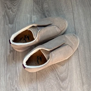 Beige/grå eytys i mocka - Stilrena slip-on sneakers från Tys i beige mocka med elastiska paneler för enkel påtagning. Skorna har vit sula, vit vadderad kant och en lila detalj baktill. Perfekta för dig som gillar minimalistisk och clean stil.