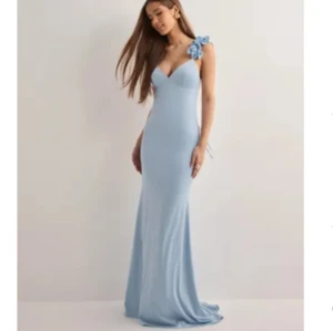 Ljusblå långklänning med blomdetalj - Säljer en elegant ljusblå långklänning med smal passform och v-ringad hals. Klänningen har en axelbandslös design med en stor blomdetalj på ena axeln som ger en unik touch. Perfekt för bal eller student, men också en fest! 👏🏼