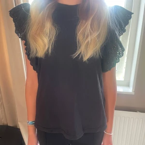 Svart blus med volangärmar - Supersnygg svart blus med volangärmar i broderad spets. Toppen har rund hals och är i mjuk bomull, perfekt för dig som vill ha något extra till outfiten. Ärmlös modell med riktigt coola detaljer på axlarna.