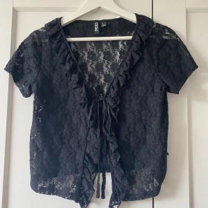 Svart spetsblus med volang från Pieces - Supersöt svart blus i spets från Pieces med kort ärm, volangkant längs v-ringningen och knyt framtill. Blommigt spetsmönster ger en romantisk vibe och toppen är lätt transparent. Perfekt att styla lager på lager eller med ett linne under. Använt en gång, nypris 340kr💕
