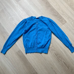 Blå kofta från Saint Tropez - Supersnygg blå kofta från Saint Tropez i storlek S. Den har rund hals, långa ärmar och knappar hela vägen framtill. Koftan är i mjukt material och har ribbade muddar vid ärmslut och nederkant. Perfekt att slänga över en t-shirt eller klänning.