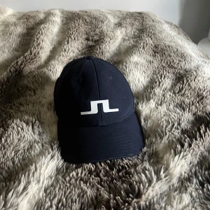 Marinblå keps från J.Lindeberg - Snygg marinblå keps från J.Lindeberg med vit logga framtill och justerbar rem baktill. Kepsen är tillverkad i lätt polyester med diskret rutstruktur och ventilerande mesh på insidan. Perfekt för dig som gillar stilren sportig look.