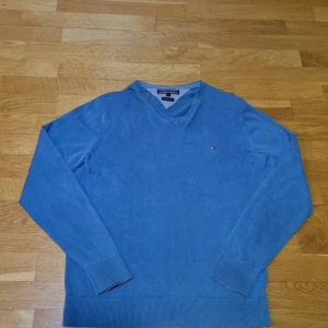 Blå v-ringad tröja från Tommy Hilfiger - Säljer en stilren blå v-ringad tröja från Tommy Hilfiger i premium bomull. Tröjan har långa ärmar och en liten logga på bröstet. Perfekt för en avslappnad men ändå snygg look.