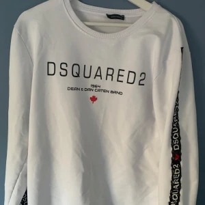Vit tröja från Dsquared2 - Säljer en stilren vit tröja från Dsquared2 med svart text och en röd lönnlövdetalj på framsidan. Tröjan har långa ärmar med ett svart band längs sidorna där märkesnamnet upprepas. Perfekt för en casual look. Obs! (Passar M och large)
