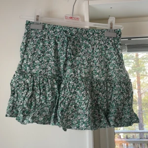 Grön blommig kjol från Zara - Säljer en söt grön blommig viskos kjol från Zara. Den har även inner shorts! Kjolen har en volangdetalj och är perfekt för varmare dagar. Den är kort och har en elastisk midja för en bekväm passform. Perfekt för att lägga till en färgglad touch till din garderob! 