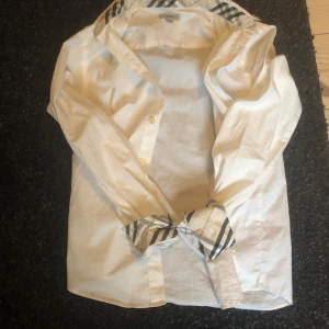 Vit skjorta från Burberry - Säljer en stilren vit skjorta från Burberry med klassiskt rutigt mönster på insidan av kragen och ärmsluten. Skjortan har långa ärmar och knappar framtill. Perfekt för en elegant look! Priset går o diskutera!