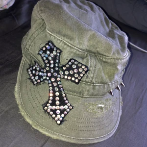 Cool y2k keps med rhinestones och nitar - Cool olivgrön keps med en glittrig korsdetalj på sidan och nitar längs kanten. Kepsen har en sliten look och är tillverkad i ett robust material. 