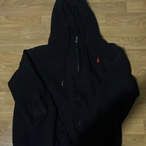 Svart hoodie från Polo Ralph Lauren - Säljer en stilren svart hoodie från Polo Ralph Lauren med dragkedja och en liten röd logga på bröstet. Perfekt för en avslappnad look. Hoodien har en klassisk passform och är tillverkad i mjukt material.