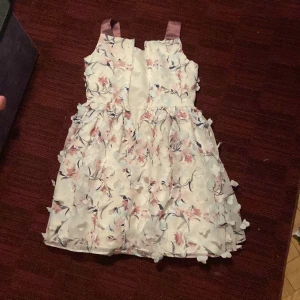 Blommig klänning med volanger - Storlek 128-130 Charmig blommig klänning med volanger och axelband i satin. Klänningen har en vit bas med rosa och blå blommönster. Perfekt för somriga tillfällen. 