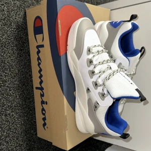 Vita sneakers från Champion - Snygga vita sneakers från Champion med blå och grå detaljer. Skorna har en robust sula med röd och svart design. De är utrustade med snörning och har en bekväm passform. Perfekta för en sportig look!