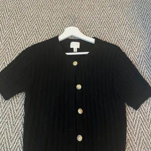 Svart ribbad kofta från H&M - Säljer en stilren svart ribbad kofta från H&M med korta ärmar och dekorativa guldknappar framtill. 
