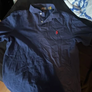 Blå pikétröja från Ralph Lauren - Säljer en klassisk blå pikétröja från Ralph Lauren med röd broderad logga på bröstet. Tröjan har korta ärmar och knappar vid kragen. Perfekt för en stilren look. Jättenajs inför sommaren. Säljer denna och en grå ifall ni går in på min profil för jag ska köpa nya kläder för jag passar inte riktigt i pikéer.