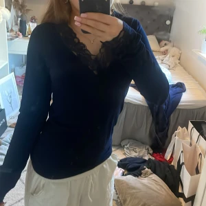 Mörkblå tröja med spetsdetaljer från RYVLS - Säljer en elegant mörkblå tröja från RYVLS med vackra spetsdetaljer vid halsringningen och ärmsluten. Tröjan har en v-ringad design och långa ärmar, perfekt för en stilren look.