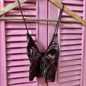 Brun satinlinne med knytning - Säljer en snygg brun satinlinne med smala axelband och knytning framtill. Perfekt för en stilren och elegant look. Passar bra till både jeans och kjol.