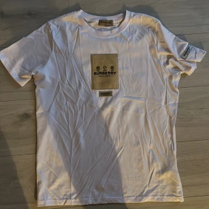 Vit t-shirt från Burberry - Säljer en stilren vit t-shirt från Burberry med ett beige tryck på bröstet och en liten etikett på ärmen. Perfekt för en avslappnad och trendig look.