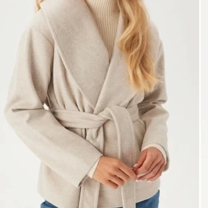 Beige kappa med bälte - Snygg beige kappa med bälte och långa ärmar. Perfekt för höst och vår. Den har en stilren design med en omlott-look och är tillverkad i ett mjukt material. Använd fåtal gånger, som helt ny! Hör av er vid frågor❤️