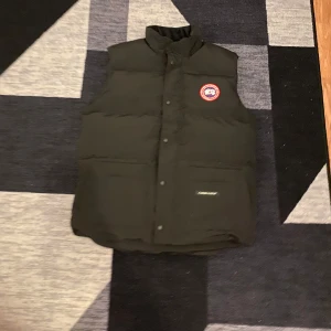 Svart dunväst från Canada Goose - Säljer en svart dunväst från Canada Goose med en klassisk design och logotyp på bröstet. Västen har en hög krage och knäppning framtill. Perfekt för kyliga dagar.