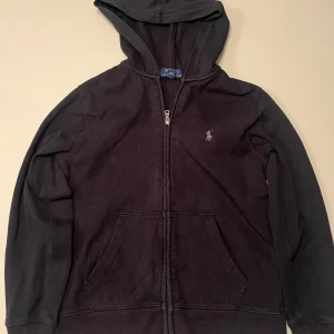 Svart hoodie från Ralph Lauren - Säljer en klassisk svart hoodie från Ralph Lauren med dragkedja och huva. Den har en liten broderad logga på bröstet och praktiska fickor framtill. Perfekt för en avslappnad stil. Storlek ca 152.