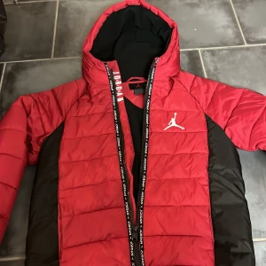 Röd och svart pufferjacka från Jordan - Säljer en snygg röd och svart pufferjacka från Jordan med ikonisk logga på bröstet. Jackan har en dragkedja med texten 'Jordan' längs med och en praktisk huva. Perfekt för kyliga dagar!