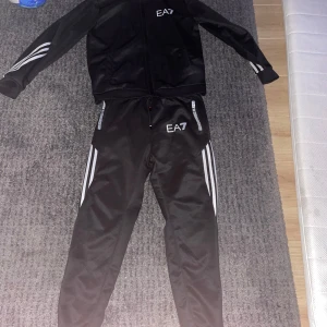 Svart träningsset från EA7 - Säljer ett svart träningsset från EA7 med vita ränder längs ärmar och ben. Setet består av en hoodie och matchande byxor, perfekt för träning eller en avslappnad dag. Hoodie med dragkedja och byxor med elastisk midja.