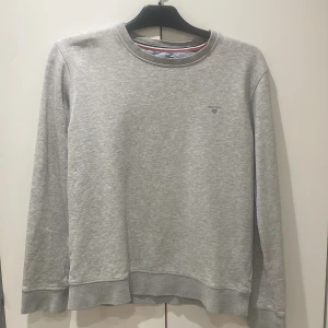 Grå sweatshirt från GANT - Säljer en stilren grå sweatshirt från GANT med broderad logga på bröstet. Tröjan har långa ärmar och rund halsringning. Perfekt för en avslappnad stil.