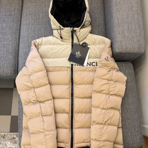 Beige pufferjacka från Moncler - Säljer en snygg beige pufferjacka från Moncler med huva och dragkedja. Jackan har en quiltad design och är perfekt för kyligare väder. Den har en stilren look med märkets logga på bröstet.