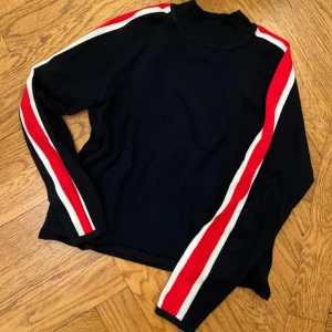 Tröja - Snygg svart tröja med långa ärmar och coola röda och vita ränder längs ärmarna. Perfekt för en sportig look eller en avslappnad dag ute. Den har en hög krage som ger extra stilpoäng.