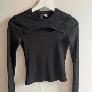 Svart ribbad topp med cut-out - Snygg svart ribbad topp från H&M med en unik cut-out detalj framtill. Toppen har långa ärmar och en tight passform som framhäver figuren. Perfekt för en trendig look!