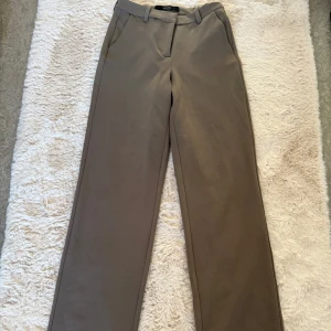 Beige kostymbyxor från Vero Moda - Snygga beige kostymbyxor från Vero Moda i en klassisk rak stil. Byxorna har en medel hög midja och är tillverkade i ett mjukt material som ger en elegant look. Perfekta för en stilren outfit.