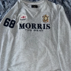 Grå tröja från Morris Gents - Säljer en stilren grå tröja från Morris Gents med broderad logga och text på bröstet. Tröjan har långa ärmar och ett klassiskt snitt. Perfekt för en avslappnad men ändå snygg look.
