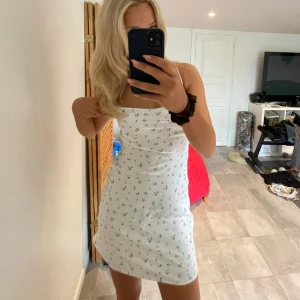 Vit blommig klänning med smala axelband - Säljer en vit satin klänning med små blå blommor och smala axelband. Klänningen har en knytning i ryggen och en kort längd som passar perfekt för varma dagar, köp via köp nu❤️