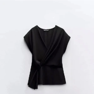 Svart omlottopp från Zara - Elegant svart omlottopp från Zara med en stilren design. Toppen har en v-ringad hals och är ärmlös, vilket ger en modern och chic look. Perfekt för att klä upp eller ner beroende på tillfälle.
