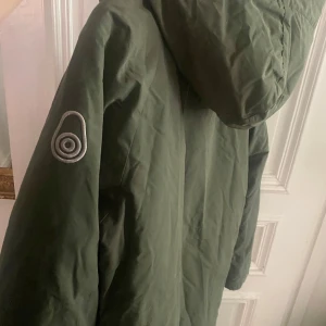 Grön parkajacka i dun från Sail Racing - Säljer en grön parkajacka från Sail Racing med huva och dragkedja. Jackan har två stora fickor framtill och en broderad logga på bröstet. Perfekt för kyligare dagar.