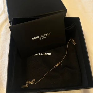 SAINT LAURENT armband - Elegant guldarmband från Saint Laurent med den ikoniska YSL-logotypen. Armbandet har en tunn kedja och en justerbar längd med en liten platta graverad med Saint Laurent Paris. Perfekt för att lägga till en touch av lyx till din stil. Nypris cirka 3500 kronor.