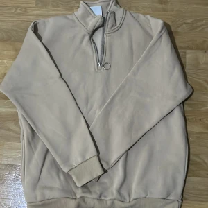 Beige tröja med dragkedja - Säljer en stilren beige tröja med dragkedja och hög krage. Tröjan har långa ärmar och ribbade muddar vid ärmslut och nederkant. Perfekt för en avslappnad look.