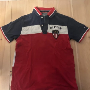 Pikétröja från Tommy Hilfiger - Säljer en snygg pikétröja från Tommy Hilfiger i blått, vitt och rött med broderad USA-sköld och Hilfiger-tryck på bröstet. Tröjan har krage med knappar och korta ärmar med röd och vit detalj.