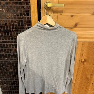 Grå polotröja i bomull - Säljer en enkel och stilren grå polotröja i mjuk bomull. Tröjan har lång ärm och en klassisk polokrage, perfekt att matcha med jeans eller kjol. Passar dig som gillar en clean och basic look.