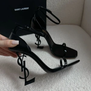 Svarta Saint Laurent klackar med logoklack - Säljer ett par svarta klackskor från Saint Laurent med öppen tå och tunn rem över foten. Klacken är formad som YSL-loggan vilket ger en unik och lyxig look. Skorna har ankelrem med spänne och är i glansigt material.