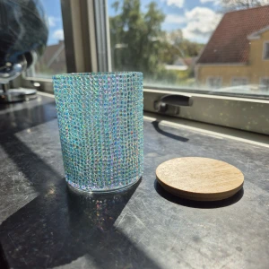 Glittrande sminkburk med trälock - En rund sminkburk handdekorerad med skimrande, ljusblå och rosa strasstenar. Burken har ett avtagbart lock i ljust trä. Perfekt för att förvara sminkborstar, pads eller små accessoarer på sminkbordet.