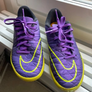 Lila Nike fotbollsskor/ innebandy med gula detaljer - Säljer ett par lila Nike fotbollsskor med gula detaljer och snörning. Skorna har en cool design med stora gula swoosh-loggor och mönster längs sidorna. Yttersulan är gul och ovandelen är i syntetmaterial. Perfekta för inomhusfotboll! 🤍Säljs pågrund av att dem kommer komma mer till användning av någon annan då dem är använd fåtal gånger🤍 Skicka meddelande för mer information/ bilder🤍 Pris kan diskuteras.