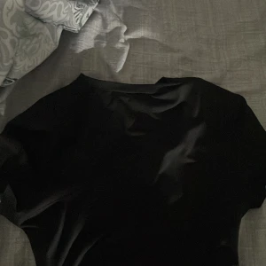 Svart topp med rynkade detaljer - Säljer en svart topp med korta ärmar och rynkade detaljer längs sidorna. Toppens material känns mjukt och stretchigt, perfekt för en stilren look. Passformen är tight och modellen är enkel men trendig.