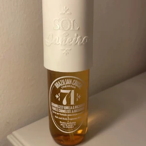 Sol de Janeiro 71 Parfym (Oanvänd) - En hår- och kroppsmist från Sol de Janeiro med doft av karamelliserad vanilj och macadamia. Flaskan är genomskinlig med gyllene vätska och har ett vitt lock med präglad logotyp. Innehåller 90 ml. Den är helt oanvänd!