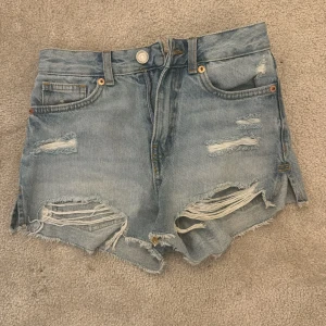 Ljusblå slitna jeansshorts från New Yorker - Ljusblå jeansshorts från New Yorker med råa kanter och flera slitningar framtill. Klassisk femficksmodell med knapp och dragkedja. Perfekta för en avslappnad och trendig look.