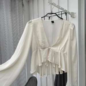 Vit blus med volang och knyt - Säljer en söt vit blus med v-ringning, volang nertill och knyt framtill. Blusen har långa, lite puffiga ärmar och är perfekt för en trendig look. Passar till både jeans och kjol.