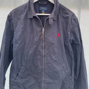 Mörkblå jacka från Polo Ralph Lauren - Säljer en klassisk mörkblå jacka från Polo Ralph Lauren med dragkedja och röd broderad logga på bröstet. Jackan har krage och två snedställda fickor framtill. Perfekt för vår och höst.